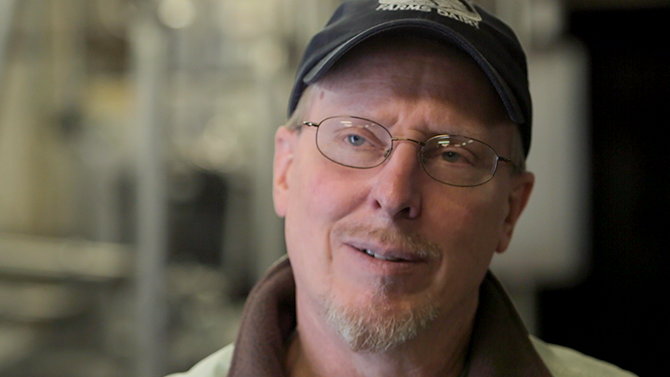 Jon Rooney a man in his fifties with a beard, glasses and hat smiles into camera, one of the team at Monument Farms Vermont Dairy Distributor