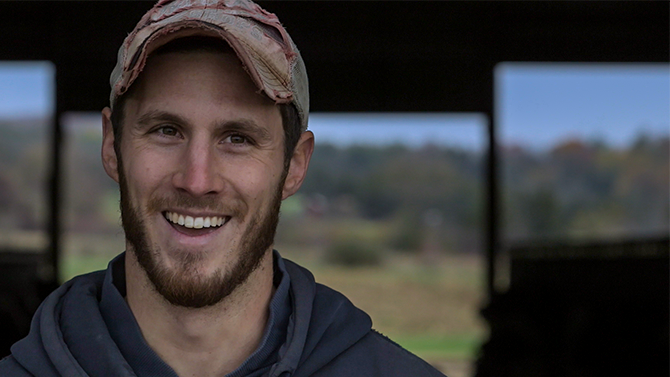 Tyler James a young man in his thirties with a beard and hat smiles into camera, one of the team at Monument Farms Vermont Dairy Distributor